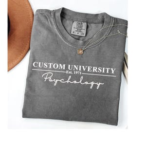 Peut inclure: Un t-shirt gris avec le texte "CUSTOM UNIVERSITY" au-dessus de "Psychology" et "Est. 1971" en blanc. Un collier pendentif doré et un chapeau marron sont également visibles.