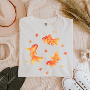 May include: A cream-colored short-sleeve t-shirt featuring a graphic print of three orange goldfish swimming among several small orange and yellow stars. The shirt has a crew neckline and a Comfort Colors brand label printed on the inside neck area.