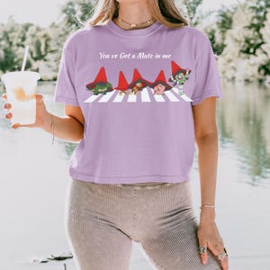 May include: A lilac-colored t-shirt with the text "You've Got a Mate in me" above a graphic of four cartoon characters crossing a white crosswalk. The characters are wearing red hats.