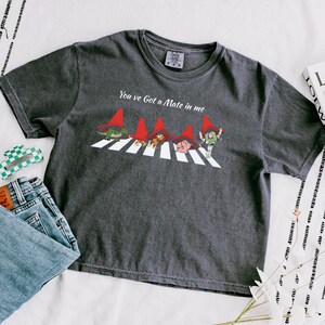 May include: Dark gray t-shirt featuring a parody of the Beatles' Abbey Road album cover. The design includes cartoon characters crossing a crosswalk, wearing red hats. The text "You've Got a Mate in me" is above the characters.