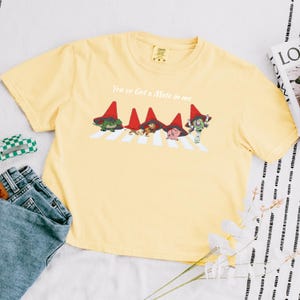 May include: A light yellow t-shirt featuring a graphic of characters crossing a crosswalk, with the text "You've Got a Mate in Me". The graphic includes characters wearing red hats. The shirt is paired with blue jeans.