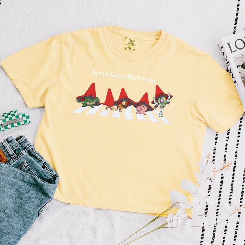 May include: A light yellow t-shirt featuring a graphic of characters crossing a crosswalk, with the text "You've Got a Mate in Me". The graphic includes characters wearing red hats. The shirt is paired with blue jeans.