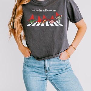 May include: Dark gray t-shirt featuring a whimsical graphic of cartoon characters crossing a street, each wearing a red hat. The text "You've Got a Mate in me" is above the graphic. The shirt is made of a soft material.