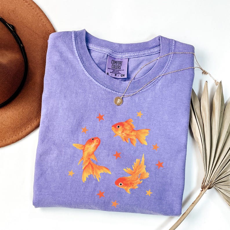 May include: A purple short-sleeved t-shirt featuring a graphic print of three orange goldfish swimming among small orange stars. The inner neck label reads Comfort Colors, 100% cotton, size S, made in Honduras. A thin gold chain necklace rests on the fabric.