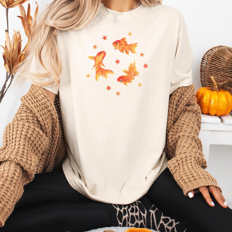 May include: A cream-colored short-sleeve t-shirt featuring a central graphic of three orange goldfish swimming in a circular formation, surrounded by small orange stars. The shirt has a crew neckline and a relaxed, loose-fitting silhouette.