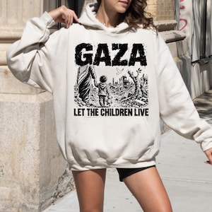 Pode incluir: Uma camisola com capuz de cor creme com uma impressão gráfica preta na frente. O design apresenta o texto GAZA no topo e LET THE CHILDREN LIVE na parte inferior, rodeando uma ilustração de uma criança a segurar um urso de peluche.