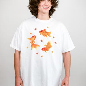 May include: A white short-sleeved t-shirt featuring a graphic print of three orange goldfish swimming in a circular formation, surrounded by small orange stars scattered around the fish. The design is centered on the chest of the garment.