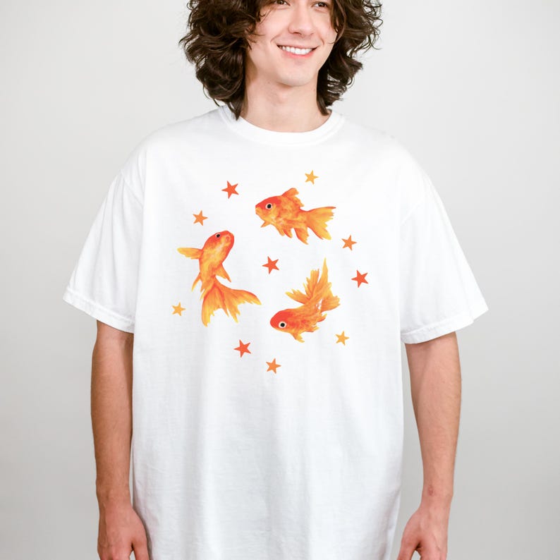 May include: A white short-sleeved t-shirt featuring a graphic print of three orange goldfish swimming in a circular formation, surrounded by small orange stars scattered around the fish. The design is centered on the chest of the garment.
