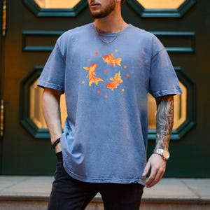 May include: A dusty blue short-sleeve t-shirt featuring a central graphic of three orange goldfish swimming in a circular formation, surrounded by small orange star shapes. The shirt has a crew neckline and a relaxed, loose-fitting silhouette.