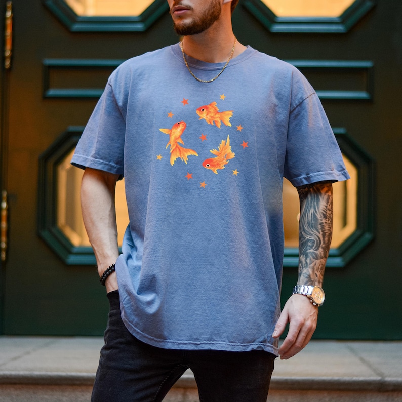 May include: A dusty blue short-sleeve t-shirt featuring a central graphic of three orange goldfish swimming in a circular formation, surrounded by small orange star shapes. The shirt has a crew neckline and a relaxed, loose-fitting silhouette.