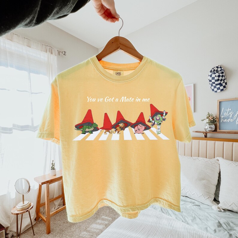 May include: Yellow t-shirt with the phrase "You've Got a Mate in Me" and a graphic of characters crossing a crosswalk. The characters are wearing red hats. The shirt is hanging on a wooden hanger.