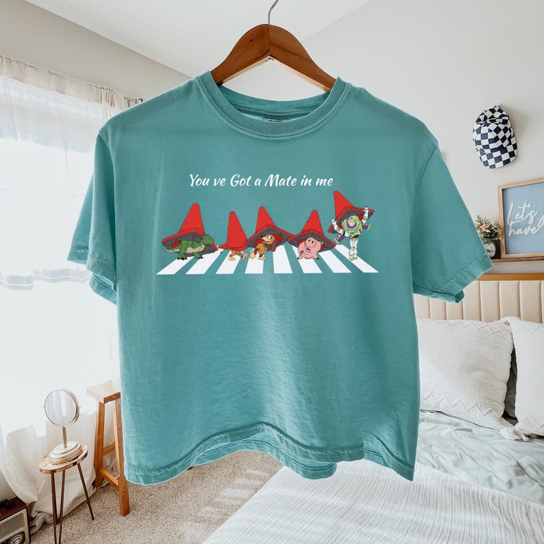 May include: Teal-colored t-shirt featuring a graphic of characters crossing a crosswalk, each wearing a red hat. The text "You've Got a Mate in me" is above the graphic. The shirt is hanging on a wooden hanger.
