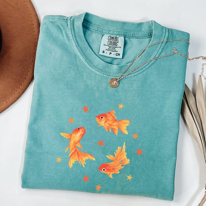 May include: A teal crew neck t-shirt featuring a graphic of three orange goldfish swimming in a circle surrounded by small orange stars. The shirt has a Comfort Colors brand label at the inner neckline indicating size S, P, CH.