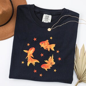 May include: A black short-sleeved t-shirt featuring a graphic print of three orange goldfish swimming among small orange stars. The shirt has a Comfort Colors brand label at the inner neckline and a crew neck collar design.
