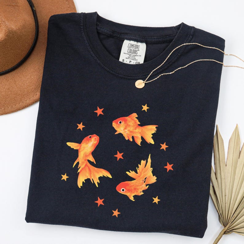 May include: A black short-sleeved t-shirt featuring a graphic print of three orange goldfish swimming among small orange stars. The shirt has a Comfort Colors brand label at the inner neckline and a crew neck collar design.