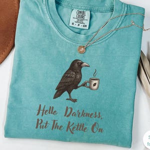 Peut inclure: T-shirt vert d'eau avec un dessin d'un corbeau tenant une tasse de café. Le texte "Hello Darkness, Put The Kettle On" est imprimé en dessous. Un collier doré avec un pendentif est posé sur le t-shirt.