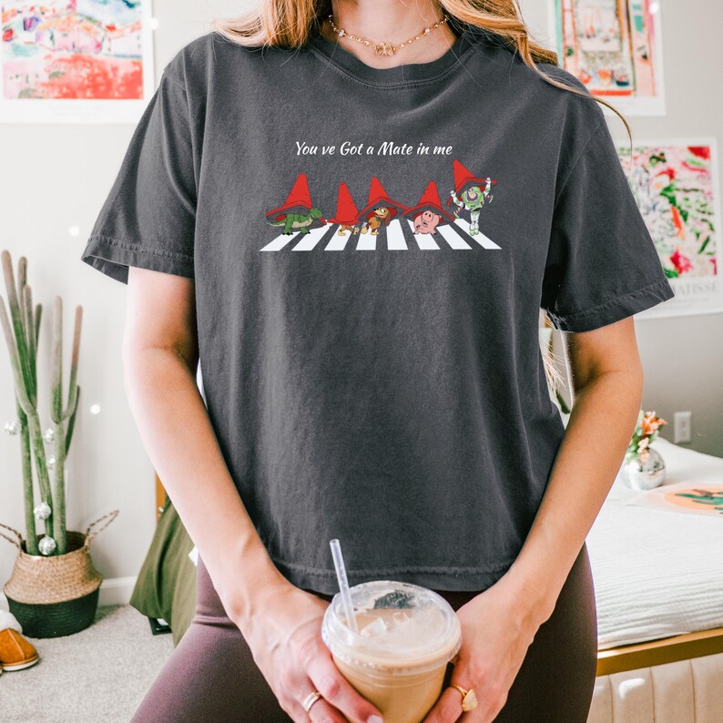 May include: Dark gray t-shirt featuring a whimsical design of gnomes crossing a crosswalk, with the text "You've Got a Mate in me" above the image. The gnomes wear red hats and are in a line. The shirt is a comfortable, casual style.