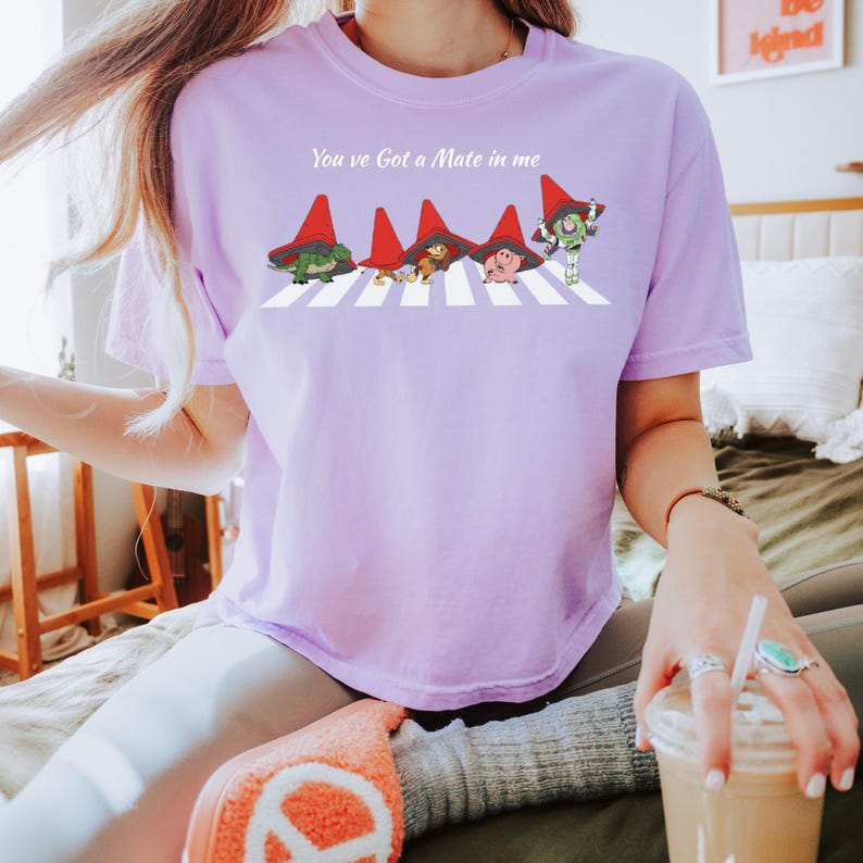 May include: A light purple t-shirt with the text "You've Got A Mate in me". The shirt features a cartoon illustration of characters crossing a crosswalk, each wearing a red hat. The characters include a dinosaur, a bear, a pig, and Buzz Lightyear.