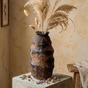 Puede incluir: Un jarrón de cerámica marrón y negro texturizado con una abertura ancha, que contiene hierba de la pampa seca y otros elementos florales secos. El jarrón está sobre un bloque de hormigón, con un fondo neutro.