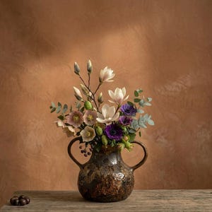 Puede incluir: Un arreglo floral en un jarrón marrón texturizado con dos asas. El ramo incluye flores de magnolia blancas, anémonas moradas y eucalipto verde. El jarrón está sobre una mesa de madera rústica.
