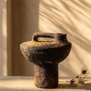 Könnte beinhalten: Eine rustikale, dunkelbraune Keramikvase mit Griff. Die Vase hat eine strukturierte Oberfläche und einen breiten, abgerundeten Boden. Der Griff ist ein horizontaler Balken. Die Vase steht auf einer Betonoberfläche.