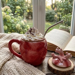 May include: A red ceramic mug filled with hot chocolate and topped with whipped cream and chocolate shavings. A stainless steel straw is in the mug. A ceramic heart-shaped object sits on a wooden coaster next to an open book.