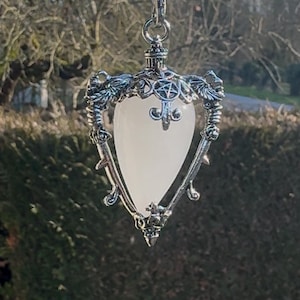 Puede incluir: Colgante plateado con una piedra translúcida en forma de corazón. El colgante cuelga de una cadena y presenta detalles ornamentales, incluyendo una flor de lis y un pentagrama. El fondo muestra un entorno natural al aire libre.