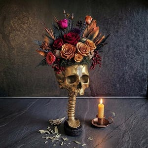 Peut inclure: Un vase doré en forme de crâne contient un arrangement floral vibrant de roses rouges, oranges et roses, de baies et de plumes. Une bougie allumée dans un porte-bougie en cuivre est à proximité. Le vase a une base en colonne vertébrale.