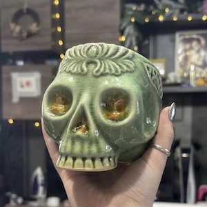 Pode incluir: Um recipiente verde em cerâmica em forma de caveira com acabamento craquelado. A caveira apresenta traços faciais detalhados e um design floral decorativo no topo. O recipiente é segurado em uma mão, mostrando seu tamanho e forma.