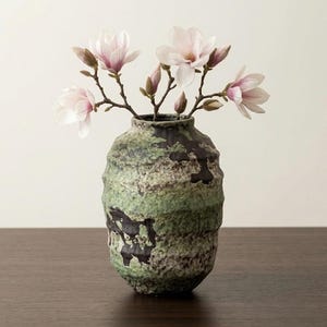 Puede incluir: Un jarrón de cerámica texturizada verde y negra con flores de magnolia rosas. El jarrón tiene una forma orgánica única y está sobre una superficie de madera oscura. Las flores están en plena floración.