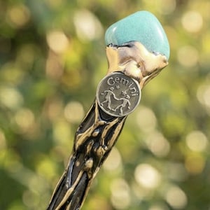 May include: A wand featuring a teal gemstone top and a bronze-toned handle with an organic, textured pattern. The handle has a small, circular emblem with the word "Gemini" and a symbol of two figures.