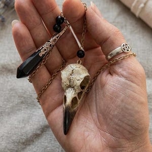 Op de afbeelding: Een hand toont een collectie gotische sieraden. Inbegrepen zijn een zwarte kristallen hanger, een vogel schedel hanger en een zilveren ring met een pentagram ontwerp. De hangers hangen aan gouden kettingen.