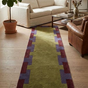 May include: A rectangular area rug featuring a solid olive green centre panel framed by a geometric border of alternating deep burgundy and muted purple rectangular blocks, creating a structured pattern that runs along the entire length of the rug.