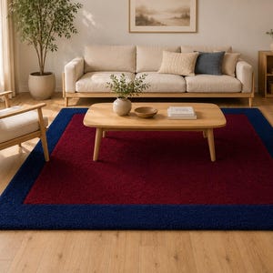 May include: A rectangular area rug featuring a deep burgundy center panel framed by a wide, solid navy blue border. The rug has a textured, plush pile surface and is positioned on a light-toned wooden floor beneath a wooden coffee table.