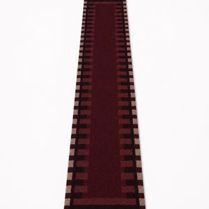 Puede incluir: Una alfombra de pasillo larga y rectangular con un panel central de color burdeos profundo sólido. El borde presenta un patrón geométrico repetitivo de bloques rectangulares que alternan entre marrón oscuro y beige claro.