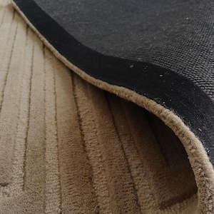 May include: Beige area rug with a geometric pattern and a black border. The rug's texture is visible, with a close-up view showing the weave and the underside of the rug.