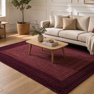 May include: A rectangular area rug featuring a braided texture with a deep burgundy center and a darker plum-colored border. The rug is crafted from natural fibers and lies flat on a light-toned wooden floor, providing a rich color contrast.
