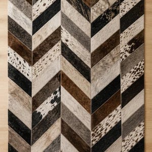 May include: A rectangular area rug featuring a chevron pattern composed of stitched cowhide strips. The design incorporates a mix of solid black, dark brown, light brown, gray, and white spotted hide textures arranged in a repeating diagonal layout.