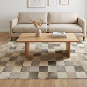 May include: A rectangular area rug featuring a patchwork design of square tiles in varying shades of beige, cream, gray, and brown. The rug is composed of individual textured pieces arranged in a grid pattern, covering the floor beneath a wooden table.