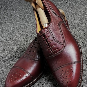 May include: Burgundy leather Oxford shoes with brogue detailing on the toe caps. The shoes have a polished finish and are laced up. Wooden shoe trees are inserted to preserve the shape. The shoes are a classic design, suitable for formal occasions.