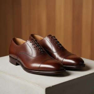 Puede incluir: Un par de zapatos de vestir de cuero marrón con diseño de puntera. Los zapatos tienen un acabado pulido, cordones marrones oscuros y están sobre una superficie clara. El fondo tiene una textura de madera cálida.