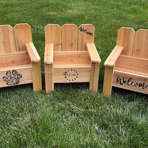 May include: Three wooden Adirondack chairs with a natural finish are arranged on green grass. One chair has a paw print design, another has "1776" with stars, and the third says "Welcome".
