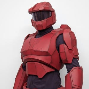 Könnte beinhalten: Rote Cosplay-Rüstung, einschließlich eines Helms mit dunklem Visier, Brustplatte, Schulterpolstern, Armschützern und Gürtel. Die Rüstung hat eine matte rote Farbe mit sichtbaren Linien und einem futuristischen Design. Das Kostüm wird über einem dunklen Hemd getragen.