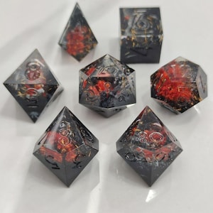 May include: A set of seven polyhedral dice with a black resin base, featuring red and gold internal designs. The dice are numbered and designed for tabletop gaming. The set includes various shapes, such as a d20, d12, and d4.