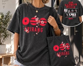 Op de afbeelding: Een antracietgrijs oversized T-shirt en bijpassende tas met rode cirkelvormige afbeeldingen, de tekst ARIRANG en roze kersenbloesemaccenten. De achterkant van het T-shirt toont de tekst BTS WORLD TOUR ARIRANG met een filmstrip van bandleden.