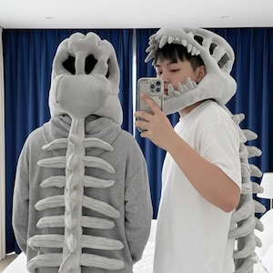 May include: Two people wearing gray dinosaur skeleton hoodies and hats. The hoodies have a spine design down the back. One person is taking a selfie with a phone. The hats are shaped like a dinosaur skull.