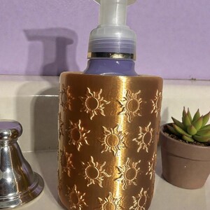 May include: A decorative soap dispenser with a gold-colored, sun-patterned sleeve. The dispenser has a clear pump and a purple bottle. A small potted succulent is visible in the background.