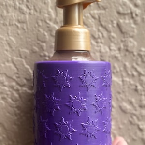 May include: A purple soap dispenser with a gold pump. The dispenser has a cylindrical shape and is decorated with embossed sun designs. The background is a textured, neutral color.
