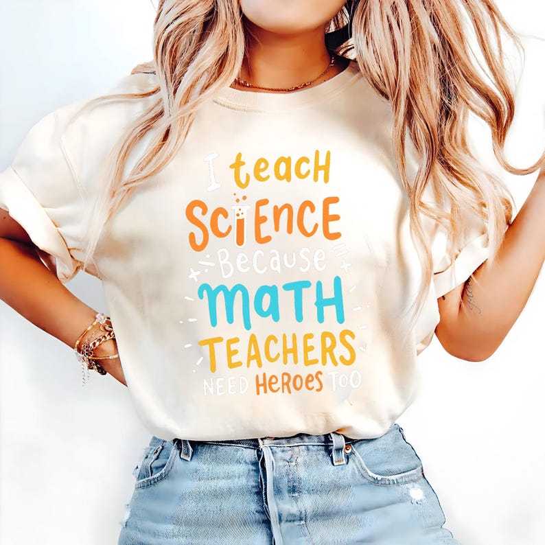May include: Cream-colored t-shirt with the text "I teach SCIENCE because MATH TEACHERS NEED HEROES TOO" in orange, blue, and yellow lettering. The shirt is paired with blue denim jeans.