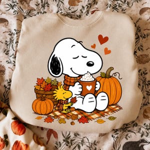 Könnte beinhalten: Ein beigefarbenes Sweatshirt mit Rundhalsausschnitt und einer Grafik von Snoopy und Woodstock. Snoopy trägt einen karierten Schal und hält eine Tasse mit einem Herz, während Woodstock daneben steht. Das Motiv zeigt Kürbisse und Herbstblätter.